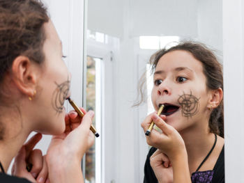 Caucasian teenage girl in a witch dress paints her lips with a black pencil looking in the mirror 