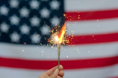 Cropped hand of person holding illuminated sparklers against american flag