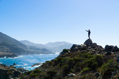 Man standing on rock by sea against clear blue sky