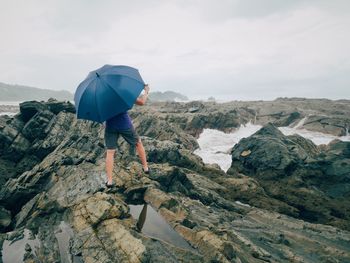 Rear view of woman with umbrella standing on rock at beach against cloudy sky