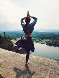 Rear view of woman doing yoga on cliff by lake against sky