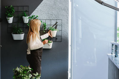 Side view of female hanging white flowerpot with green plant on gray wall with various flowers in light room