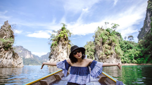 Portrait of young woman pouting while sitting on boat in sea against rock formations and sky