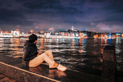 Woman sitting on river with illuminated city in background