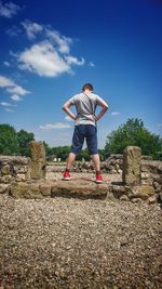 Rear view of boy with hands on hip standing on stone against sky