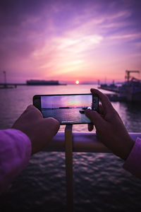 Hand holding smart phone by sea against sky during sunset