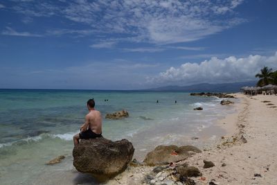 Rear view of shirtless man sitting on rock at beach against sky