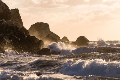 Waves splashing on rocks at shore against sky