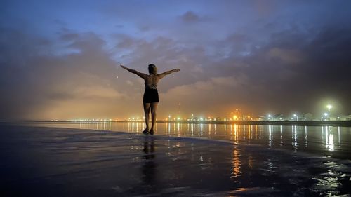 Rear view of woman with arms outstretched standing at shore against sky during sunset