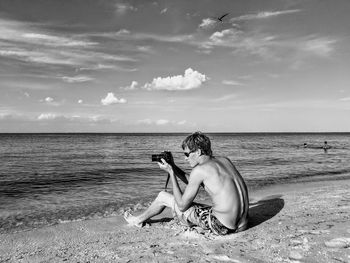 Shirtless man photographing while sitting on shore at beach against sky