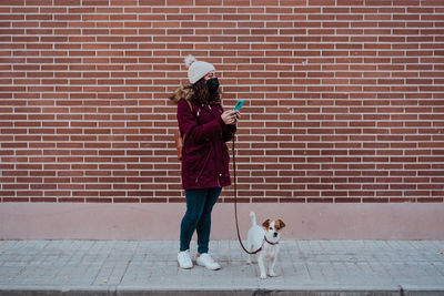 Woman with dog using phone while standing against brick wall