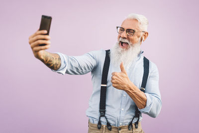 Cheerful bearded senior man taking selfie with mobile phone against purple background