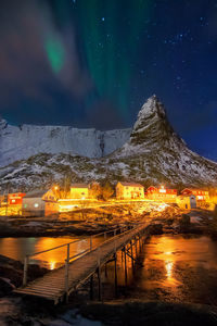 Wooden bridge over river leading towards illuminated houses and mountain at winter