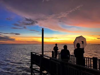 Silhouette men with umbrella on pier over sea against sky during sunset