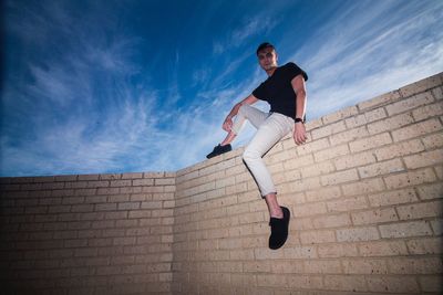 Low angle view of man sitting on retaining wall against sky
