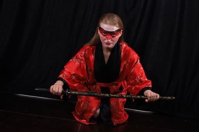 Portrait of young female model holding samurai sword while kneeling against black curtain