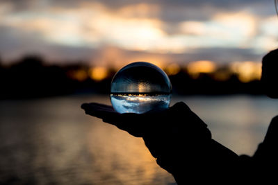 Silhouette hand holding crystal ball against lake during sunset