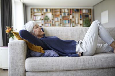 Smiling senior woman with hands behind head lying down on sofa at home