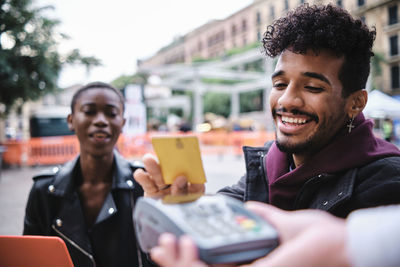 Smiling man paying through card whiles sitting with friend at sidewalk cafe