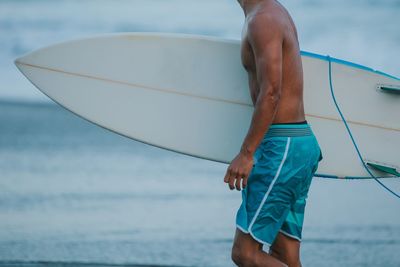 Midsection of man holding surfboard while walking against sea at beach