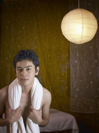 Portrait of shirtless young man with towel sitting in bedroom