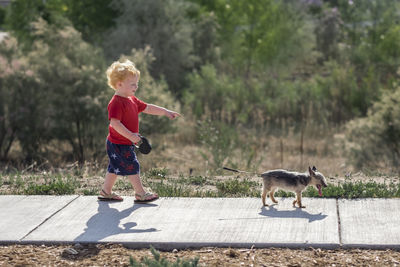 Side view of cute baby boy pointing while walking with puppy on footpath in park