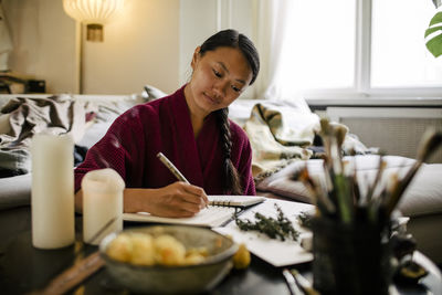 Smiling woman writing in book while sitting with candles and herbs at table