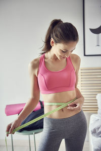Young woman measuring stomach with tape measure in bedroom at home