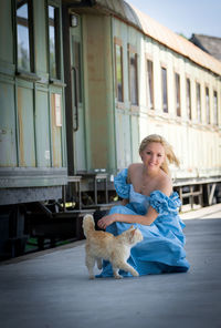 Portrait of beautiful model in blue evening gown crouching by cat at railroad station platform