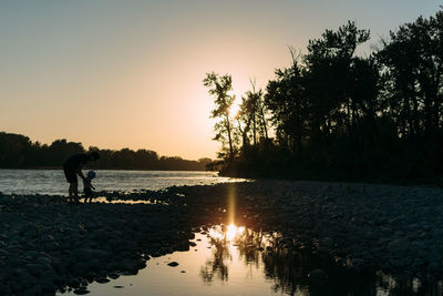 Silhouette man with son standing on riverbank against sky during sunset