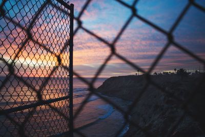 Scenic view of sea against sky during sunset seen through chainlink fence