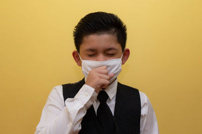 Boy wearing mask coughing while standing against yellow wall