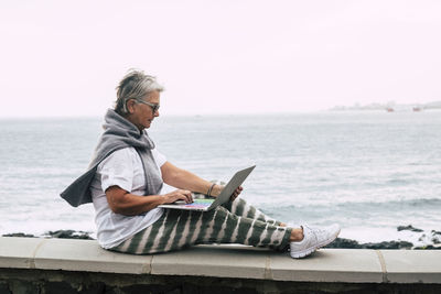 Senior woman sitting on wall by the sea, using laptop