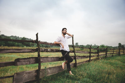 Full length of young man standing by fence on grassy field against sky