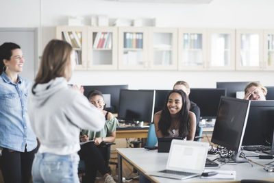 Teenage high school student standing by smiling teacher in front of friends sitting in computer lab