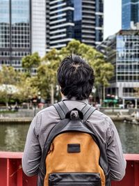 Rear view of man standing at metal fence and looking at river and skyscrapers in the city.