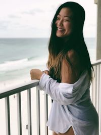 Smiling young woman standing by railing against sea