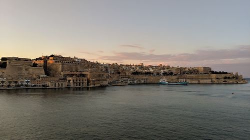 Buildings by sea against sky during sunset in city