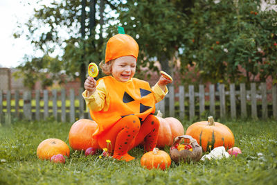 Full length of cute girl wearing costume sitting by pumpkin at park