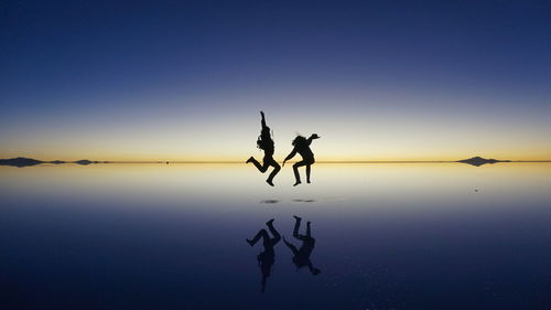 Silhouette people jumping at lake against sky during sunset