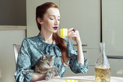 Beautiful elegant woman with a cat sits in the kitchen and holds a teacup with wine