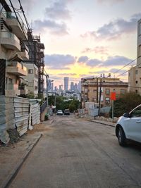 Cars on road by buildings against sky during sunset