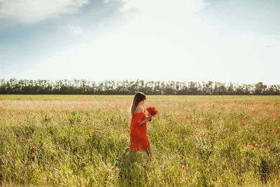 A girl in a red dress above the knee stands in a poppy field in the afternoon