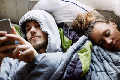 High angle view of young man using smart phone while lying by friend on bed at home