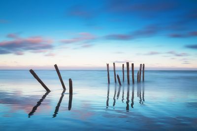 Wooden posts in sea against sky at sunset