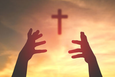 Cropped image of silhouette hands catching cross against sky during sunset