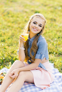 Young happy woman holds a glass of orange juice in her hand while sitting on a picnic blanket