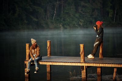 Woman photographing female friend sitting on pier in lake