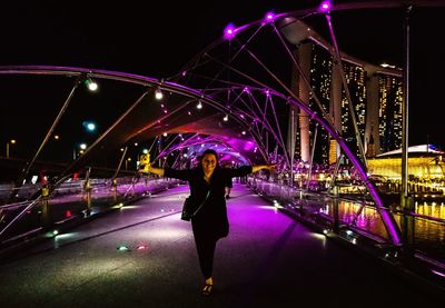 Full length of smiling woman with arms outstretched standing on illuminated bridge at night