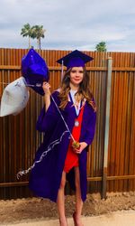 Portrait of smiling young woman wearing graduation gown standing at backyard